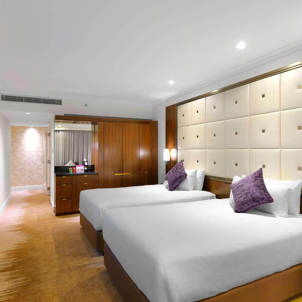 Amora Hotel Jamison Sydney Pokoj fotografie The photo shows a modern hotel room featuring two twin beds with white bedding and purple decorative pillows. The walls are designed with soft beige panels, creating a luxurious ambiance. There’s wooden furniture, including a cabinet against one wall