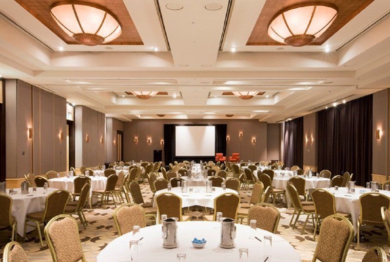 Amora Hotel Jamison Sydney Exteriér fotografie The photo shows a large banquet or conference room. There are several round tables arranged throughout the space, each covered with white tablecloths. The tables are set with water pitchers and glasses. In the center of the room, there is a projectio